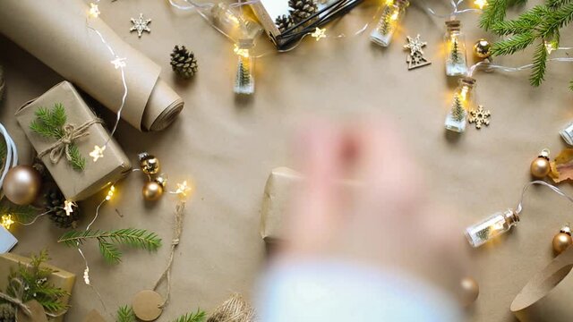 Hands pack a gift for Christmas and new year in eco-friendly materials: kraft paper, live fir branches, cones, twine, tied with a bow. Tags with mock up, natural decor, hand made, flatly. Festive mood