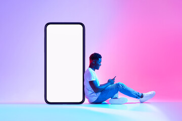 Young black man sitting next to huge smartphone with empty screen, advertising new mobile application or website, mockup
