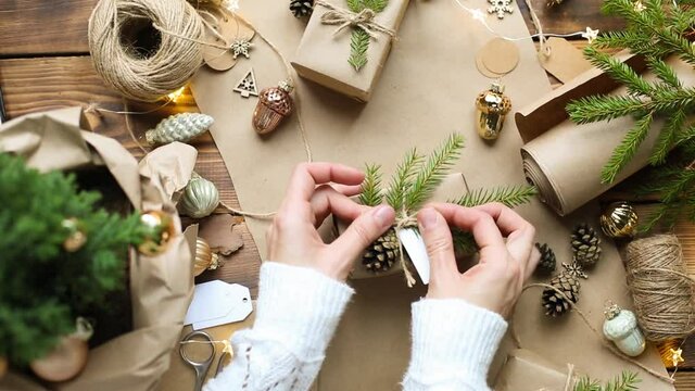 Hands pack a gift for Christmas and new year in eco-friendly materials: kraft paper, live fir branches, cones, twine, tied with a bow. Tags with mock up, natural decor, hand made, flatly. Festive mood
