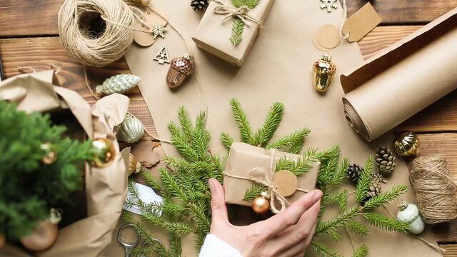 Hands pack a gift for Christmas and new year in eco-friendly materials: kraft paper, live fir branches, cones, twine, tied with a bow. Tags with mock up, natural decor, hand made, flatly. Festive mood