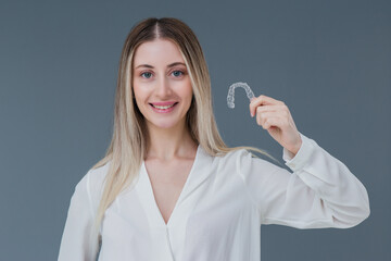 Beautiful smiling blonde woman is holding an invisible teeth aligner for orthodontic purposes. Includes copy space.