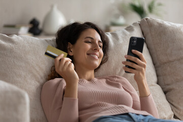 Close up smiling woman holding gold credit card and smartphone, relaxing on cozy couch at home,...