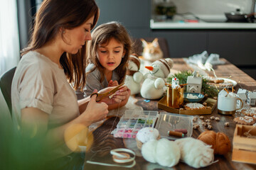 Little cute girl and young beautiful woman make knitted snowmen. Mom and daughter are sewing on...