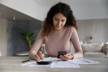 Smiling woman using smartphone and calculator, working with financial documents, receipts, young...
