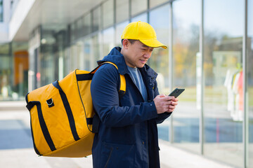 asian male food courier using mobile using mobile phone app on smartphone while standing city street with insulated with thermal backpack delivering food. looking customer address. purchase online map