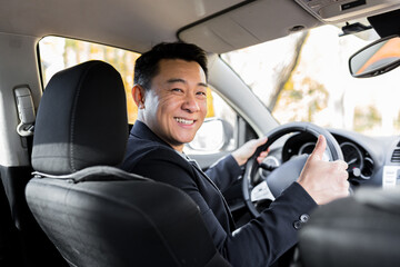 A man driving an Asian car behind the wheel looks at the camera and smiles