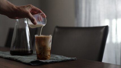man pours milk into fresh coffee over in glass on wood table