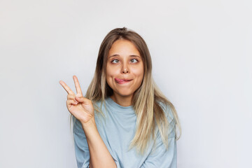 A young pretty caucasian ridiculous blonde woman in an oversize t-shirt shows tongue and a peace gesture with her hand grimacing and showing a funny face isolated on a white background