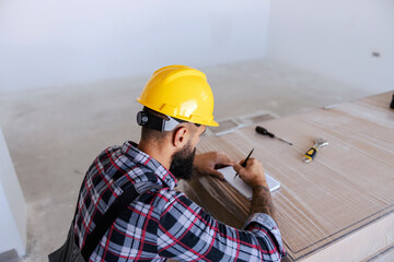 A rearview of bearded carpenter crouches in new apartment and calculating materials for wooden floor. He is writing down if it is enough materials for the entire apartment.