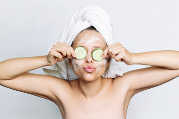 A young funny woman with a white towel on her head after a shower holds cucumber slices, covers her eyes making a face mask, sending an air kiss isolated on a gray background. Skin care, cosmetology