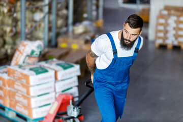 A hardworking bearded warehouse worker is pulling a forklift full of freight and relocating it to...