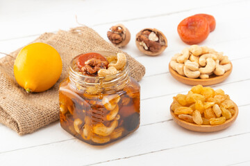 Mixture of walnuts, cashews, raisins, dried apricots and prunes with honey in a glass jar next to lemon on burlap on a white wooden table