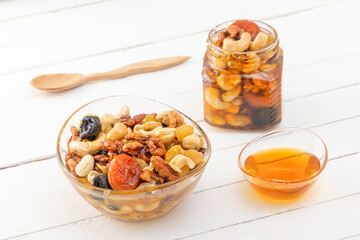 Glass cup and jar filled with a mixture of walnuts, cashews, raisins, dried apricots and prunes with honey all together with a wooden spoon on a white board table