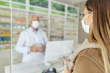 Obraz premium Buying and selling prescription drugs and pharmacist advice. A male pharmacist blurred in the background behind the counter selling drugs to an adult girl wearing a protective mask during corona virus