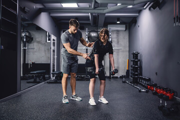 Fit woman in electric muscular suit to stimulate exercise at training center. A male trainer helps her improve her body position. A young woman in an EMS suit holds dumbbells in her hands