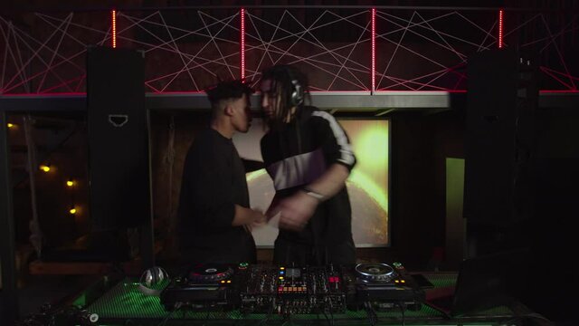Positive stylish black male clubber greeting, embracing and asking cool middle eastern dj to play techno dance music while musician mixing tracks using soundboard and cd player turntable in club.