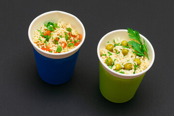 Prepared Instant Noodles with Green Pea, Carrot and Greens in Blue and Green Cups on Black Background. Quick Lunch or Unhealthy Fast Food