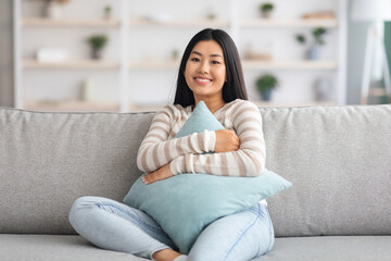 Young Asian Woman Sitting On Couch, Cuddling Pillow And Smiling At Camera