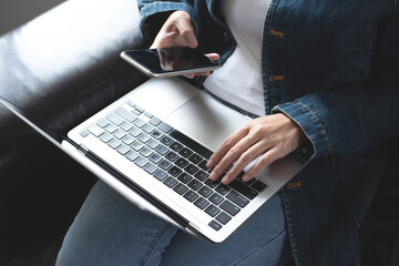 Young casual business woman, freelancer in blue jeans and sweater sitting on sofa using mobile...