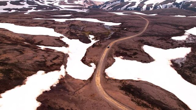 Revolving aerial shot in Iceland of car approaching snow covered mountains in highlands. Dirt road, epic snowy landscape. 