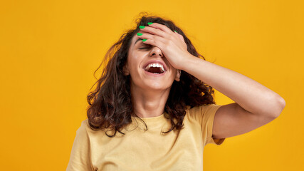Portrait of woman with curly dark hair forgetting something, slapping forehead with palm and closing eyes isolated over yellow background - obrazy, fototapety, plakaty