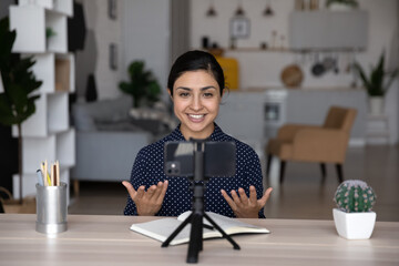Confident smiling young Indian ethnicity female blogger recording educational video on cellphone standing on stabilizer, sharing professional knowledge, streaming live stories online, sitting at table - obrazy, fototapety, plakaty