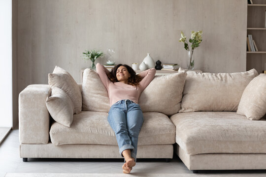Smiling barefoot woman resting on comfortable couch at home alone, happy beautiful young female with closed eyes stretching, daydreaming or taking day nap, enjoying leisure time, no stress concept