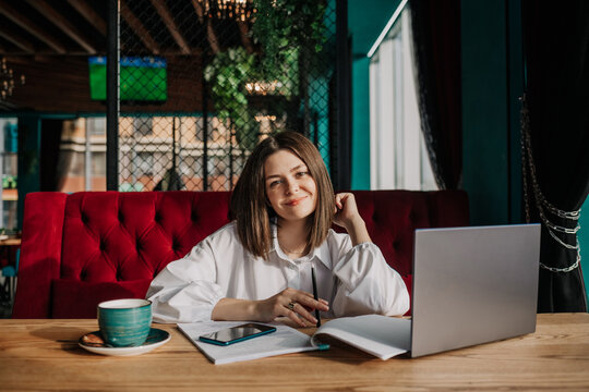 a happy brunette business woman in a white shirt is sitting at a table in a cafe with a laptop