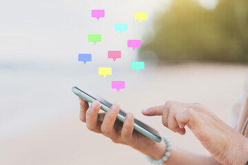 Woman hand using smart phone with message chat icon flying on tropical beach abstract background. Technology business and travel vacation concept.