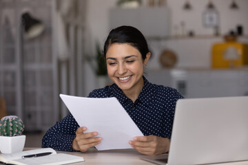 Happy young Indian businesswoman reading paper letter, feeling excited with good news in...