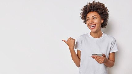 Waist up shot of cheerful woman smiles broadly holds mobile phone sends text message points thumb away on left side copy space for your advertising content. People technology advertising concept