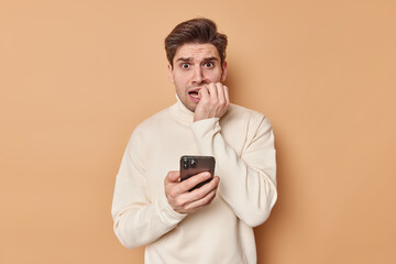 Anxious adult man bites finger nails reads chat nervously holds mobile phone wears casual jumper...
