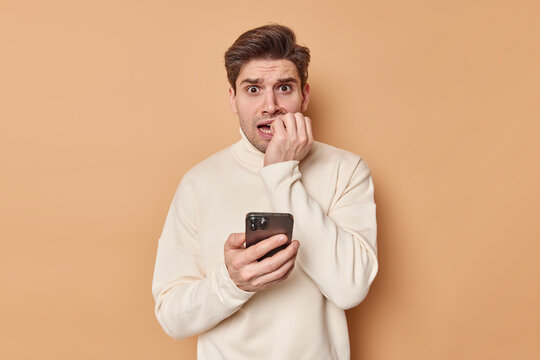 Anxious adult man bites finger nails reads chat nervously holds mobile phone wears casual jumper has scared expression made huge mistake isolated over beige background. What should I do help