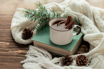 Mug of warming drink with cinnamon sticks on a book on a background of fir branches and cones. Knitted white scarf envelops a mug of hot mulled wine with spices on a wooden background. Cozy winter