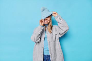 Playful energetic young European woman enjoys life hides behind hat wears stylish grey jacket has...
