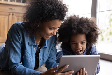 Caring smiling young African American mother or nanny teaching small multiracial child girl using digital tablet, lying together on warm heated floor, watching funny cartoons online, tech addiction.