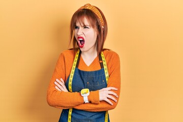 Redhead young woman dressmaker designer wearing atelier apron angry and mad screaming frustrated and furious, shouting with anger. rage and aggressive concept.