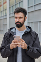 Photo of thoughtful handsome man dressed in black anorak uses mobile phone for chatting online...