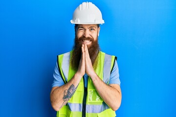 Redhead man with long beard wearing safety helmet and reflective jacket praying with hands together asking for forgiveness smiling confident.