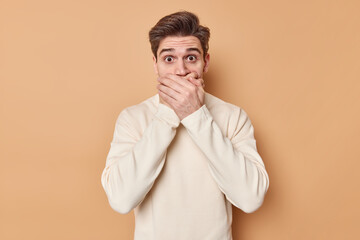 Portrait of impressed scared man slaps mouth with palms stares startled at camera hears bad atonishing news wears casual jumper isolated over beige background stands in awe. Reactions concept