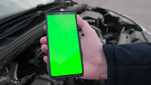 Male hand shows a smartphone with a green screen on the background of the engine and auto parts under the hood
