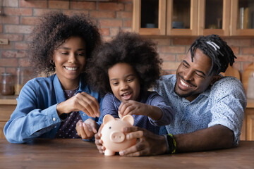 Caring young African American couple parents teaching small biracial kid daughter saving money,...