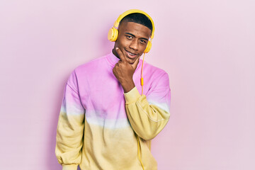Fototapeta premium Young black man listening to music wearing headphones looking confident at the camera smiling with crossed arms and hand raised on chin. thinking positive.