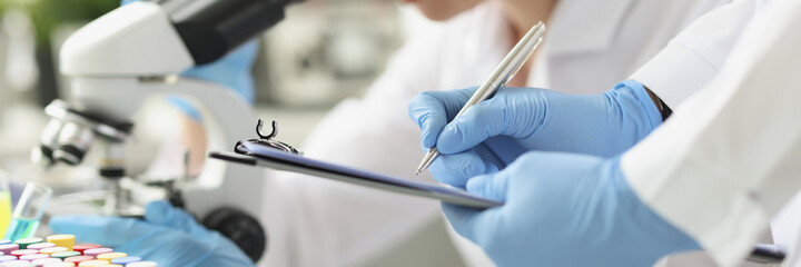 Woman chemist looking through microscope in laboratory. Assistant writing data to paper