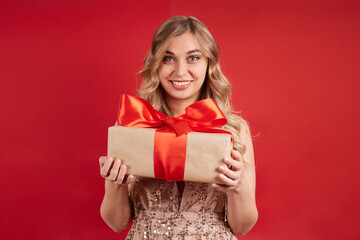 beautiful woman in a smart dress, holding a gift with her hands, smiling, looking at the camera on a red background.