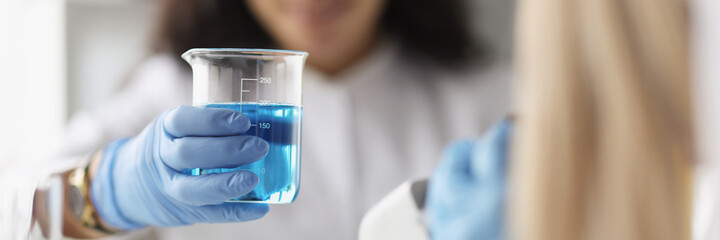 Woman chemist holding glass beaker with blue liquid in front of microscope in laboratory