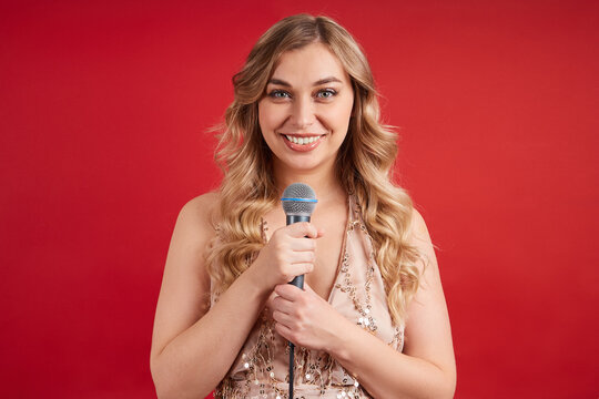 Charming blonde in a beautiful dress with a microphone posing on a red background