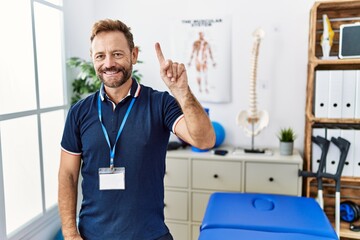 Middle age physiotherapist man working at pain recovery clinic showing and pointing up with finger number one while smiling confident and happy.