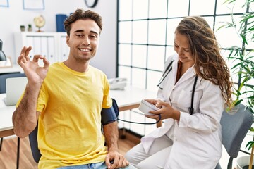 Young doctor woman checking blood pressure on patient doing ok sign with fingers, smiling friendly...