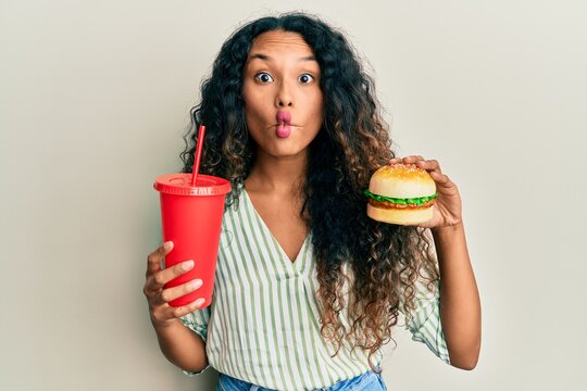 Young latin woman eating a tasty classic burger and soda making fish face with mouth and squinting eyes, crazy and comical.
